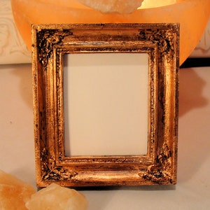 May include: A small, square, ornate gold picture frame with a blank white center. The frame has detailed carvings and a distressed finish. The frame is likely for displaying a small photograph or artwork. The frame is sitting on a white surface.