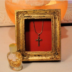May include: A gold-framed picture with a red background and a gold cross necklace inside. The frame has ornate details and is sitting on a white surface.