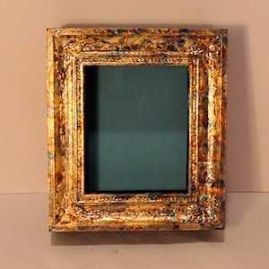 May include: A small, ornate picture frame with a gold-colored, textured finish. The frame has a rectangular opening with a teal-colored background. The frame's design includes intricate details and a vintage aesthetic.