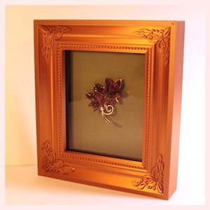 May include: A framed vintage brooch with a floral design. The brooch features dark red petals and a silver-colored stem. The gold-colored frame has ornate corner details and a dark green background.