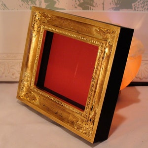Puede incluir: Un marco de imagen de pan de oro con un respaldo negro y un passe-partout rojo. El marco tiene un dise&ntilde;o decorativo y ornamentado.
