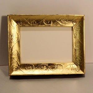 May include: A rectangular gold picture frame with ornate floral detailing. The frame has a bright, metallic finish and a white interior, perfect for displaying a photograph or artwork. The frame is made of a sturdy material.