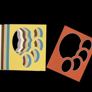 May include: A stack of square paper mats in yellow, blue, brown, white, and lavender, each featuring a large circular cutout and three smaller oval cutouts. Beside the stack is a single orange square mat with identical circular and oval cutouts.