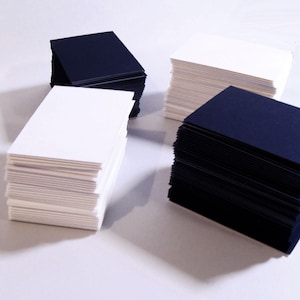 May include: Stacks of white and navy blue rectangular cards are arranged on a white surface. The cards are neatly stacked, with the edges of each card visible. The image is well-lit, highlighting the texture and color of the cards. The cards appear to be business cards or similar stationery.