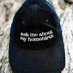 May include: Black corduroy baseball cap with the text "ask me about my homebirth" embroidered in white. The cap has a curved brim and a structured crown. The background is a blurred view of tree bark.