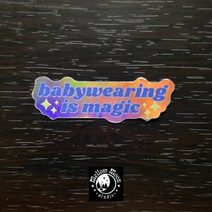 May include: A holographic sticker with the text "babywearing is magic" in a blue font with a rainbow background. The sticker has two small stars on either side of the text. The sticker is from Mellow Moon Studio.