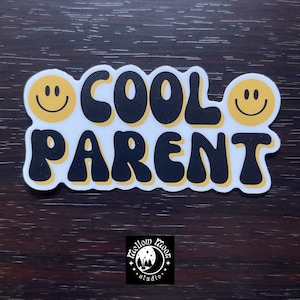 May include: A white sticker with the words "COOL PARENT" in black, retro-style font. Yellow smiley faces are on each side of the text. The sticker has a yellow outline and a small logo at the bottom.