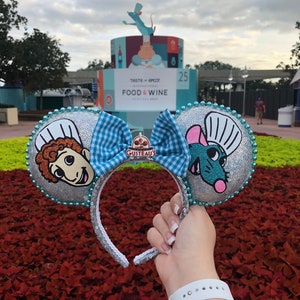 May include: A pair of silver glitter Minnie Mouse ears with blue and white checkered bow. The ears feature a chef and a rat from the movie Ratatouille. The ears are held in front of a sign that says "Taste of Epcot International Food & Wine Festival".
