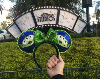 May include: A pair of Disney-themed ear headbands featuring the Little Green Men from Toy Story. The ears are blue with green sequin trim and the bow is green. The background includes the Disney's Hollywood Studios sign.