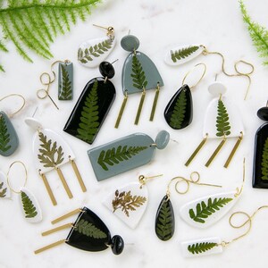 May include: A collection of handmade earrings featuring real fern leaves encased in resin. The earrings come in various shapes, including teardrops, rectangles, and geometric designs, with gold-colored metal accents. Colors include white, black, and muted green.