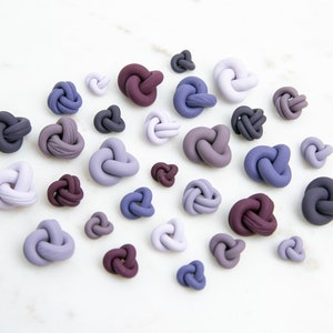 May include: A collection of 25 small, knotted clay ornaments in various shades of purple, gray, and brown. The ornaments are arranged on a white surface.