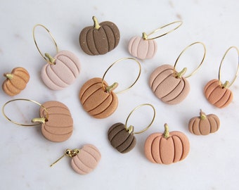 May include: A collection of pumpkin-shaped earrings in various shades of brown, tan, and pink. Each pumpkin has a gold-colored stem and is attached to a gold hoop earring. The earrings are arranged on a white surface.