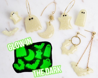 May include: A selection of glow-in-the-dark Halloween earrings featuring ghost, bat, and crescent moon designs. The earrings are pale yellow with glitter and gold-coloured metal findings. The text "GLOW IN THE DARK" is also visible in the image.