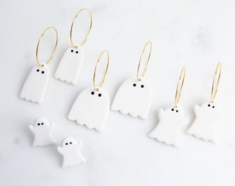 May include: A set of white ceramic ghost earrings and studs. The earrings are hoop style with a white ghost charm hanging from each hoop. The studs are small and feature a white ghost charm. The ghosts have black eyes.