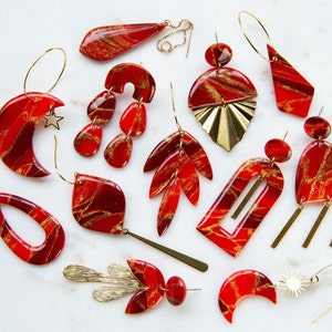May include: A collection of red and gold marbled earrings with various shapes and designs, including crescent moons, leaves, and geometric shapes.
