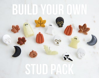 May include: A selection of Halloween-themed stud earrings featuring ghosts, pumpkins, leaves, skulls, bats, and moons. The earrings are in orange, black, white, and gold. The text "BUILD YOUR OWN STUD PACK" is displayed in white.
