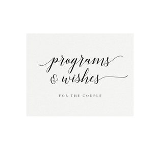 May include: A rectangular card with the words "programs & wishes" in elegant black script. Below, the text "FOR THE COUPLE" is printed in a smaller, simple font. The card has a textured, off-white background.