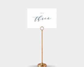 May include: A gold-colored table number holder with a round base and a slender stem. The holder displays a white card with the word "three" in elegant navy blue script. The card is held in place by a small ring at the top.