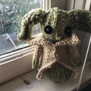 May include: A green knitted plush toy with large ears and black button eyes. The toy is wearing a tan knitted waistcoat.