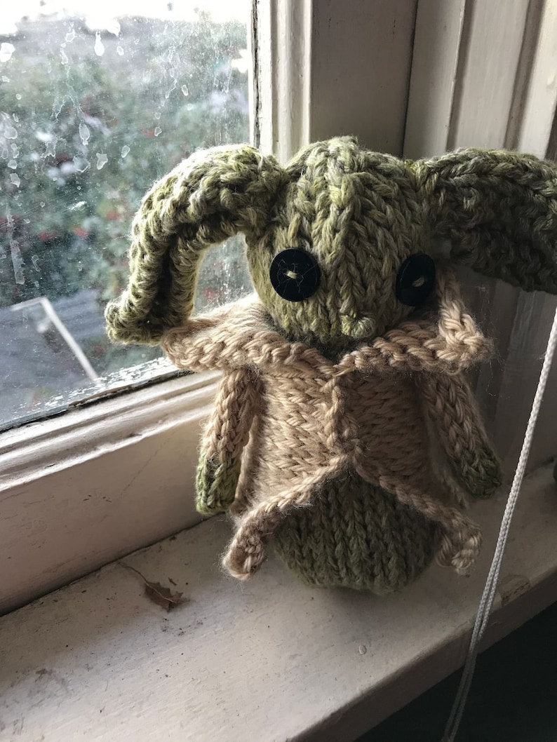 May include: A green knitted plush toy with large ears and black button eyes. The toy is wearing a tan knitted waistcoat.