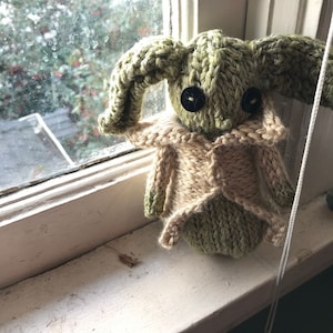 May include: A green knitted plush toy with large ears and black button eyes. The toy is wearing a tan knitted waistcoat and is sitting on a windowsill.