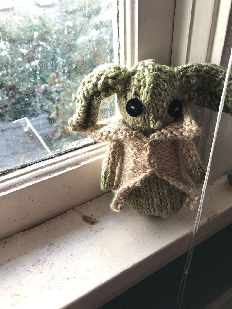May include: A green knitted plush toy with large ears and black button eyes. The toy is wearing a tan knitted waistcoat and is sitting on a windowsill.