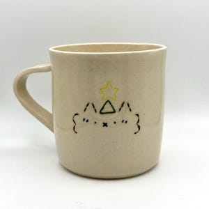 May include: A white ceramic mug with a hand-painted design of a stylised cat with a yellow star on top of a green triangle.