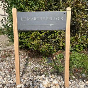 May include: A wooden signpost with a black slate sign that reads "LE MARCHE SELLIOIS" with an arrow pointing to the right.