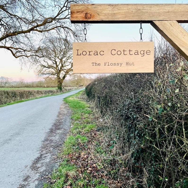 May include: A wooden sign with the text "Lorac Cottage The Flossy Hut" hanging from a wooden post. The sign is in front of a country road and a hedge.