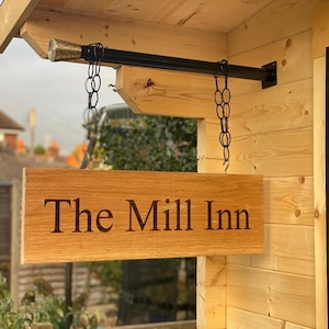 May include: A wooden sign with the text "The Mill Inn" hanging from a black metal rod with chains. The sign is attached to a wooden building.