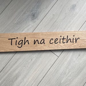 以下が含まれることがあります： 黒い筆記体で「Tigh na ceithir」と書かれた長方形の木製サイン。サインは明るい色の木材でできており、明るい灰色の木製の表面に置かれています。