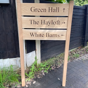 May include: A wooden signpost with three lines of text: Green Hall ↑, The Hayloft →, White Barns ←. The signpost is made of natural wood and has a rustic look.