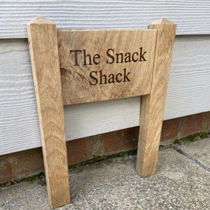 May include: A wooden sign with the text "The Snack Shack" engraved on it. The sign is attached to two wooden posts.