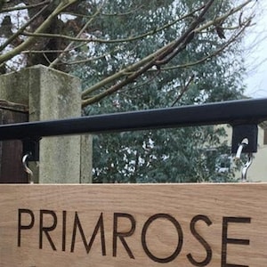 以下が含まれることがあります： 「PRIMROSE」と刻まれた木製の看板。看板は木製のフェンスに取り付けられた黒い金属棒から吊り下げられている。