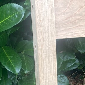May include: Close-up of a light wood post with a square top and two holes drilled into the side. The post is surrounded by green leaves.