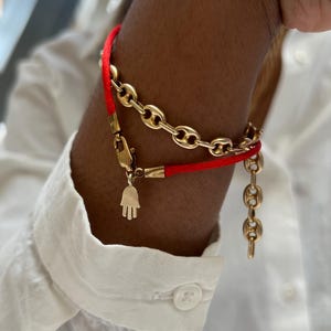 Può includere: Braccialetti a catena dorati e un braccialetto con cordino rosso. Un braccialetto presenta un piccolo ciondolo a forma di mano dorata. I braccialetti sono indossati sul polso di una persona che indossa una camicia bianca abbottonata.