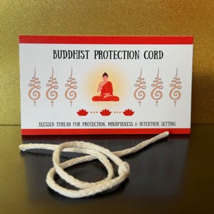 May include: A white, off-white, and red rectangular card featuring the text BUDDHIST PROTECTION CORD, an illustration of a seated figure in red, and six tan symbols. Below the card lies a single, coiled, off-white, braided cotton cord.