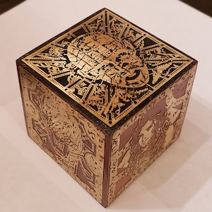 May include: A wooden box with a gold-colored design on all sides. The top of the box features a detailed, intricate design of a face.