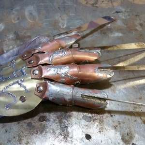 May include: A handcrafted metal glove with four long, sharp blades. The glove has a leather wrist strap and is made from copper, brass, and silver-coloured metal. The blades are approximately 15 cm long, and the design has a weathered appearance.