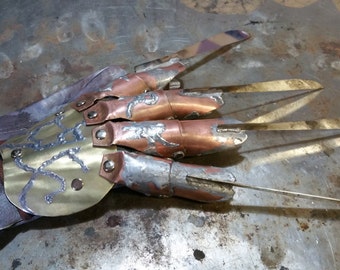 May include: A handmade metal glove with four long, sharp blades. The glove features a leather wrist strap and is constructed with copper, brass, and silver-colored metal. The blades are approximately 6 inches long, and the overall design has a weathered appearance.