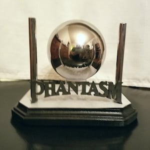 May include: A chrome sphere sits between two vertical metal rods, mounted on a white and black base. The word "PHANTASM" is displayed in dark letters on the base. The sphere reflects the surrounding environment.