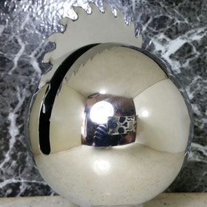 May include: A silver, round, decorative ornament with a saw blade design on top. The ornament is reflective and shows a reflection of a person holding a skull-shaped object.