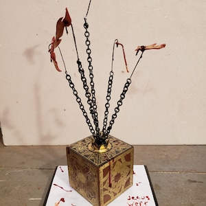 Puede incluir: Un cubo de color dorado con diseños intrincados y cadenas unidas a dedos ensangrentados, sentado sobre una base negra con las palabras "Jesús lloró" escritas en rojo.
