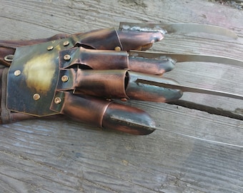 May include: A brown leather glove with copper-colored metal plating and four long, sharp, silver blades extending from the fingertips. The metal has a distressed, aged appearance, with visible rivets.