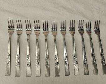 May include: A set of eleven silver-toned metal forks arranged in a row. Each fork features a four-tine head and a flat handle decorated with an embossed floral pattern consisting of a stem with leaves and a single rose bloom at the base.