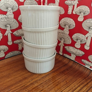 May include: A stack of four white ceramic ramekins with vertical fluted sides. Each ramekin features a smooth, glossy finish and a consistent cylindrical shape, designed for individual servings of baked goods or savoury dishes in a kitchen setting.