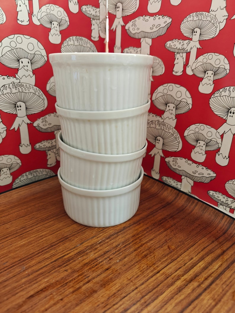 May include: A stack of four white ceramic ramekins with vertical fluted sides. Each ramekin features a smooth, glossy finish and a consistent cylindrical shape, designed for individual servings of baked goods or savoury dishes in a kitchen setting.