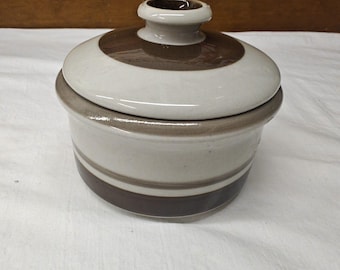May include: A round ceramic lidded container featuring a glossy off-white finish with horizontal bands of dark brown and light brown glaze. The lid includes a central knob handle, and the base has a matching brown band around the bottom edge.