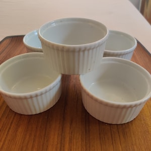 May include: A collection of five white, fluted ramekins arranged on a wooden cutting board. The ramekins are round with a classic design, perfect for baking and serving individual portions of desserts or savory dishes. The background includes a table and a window.
