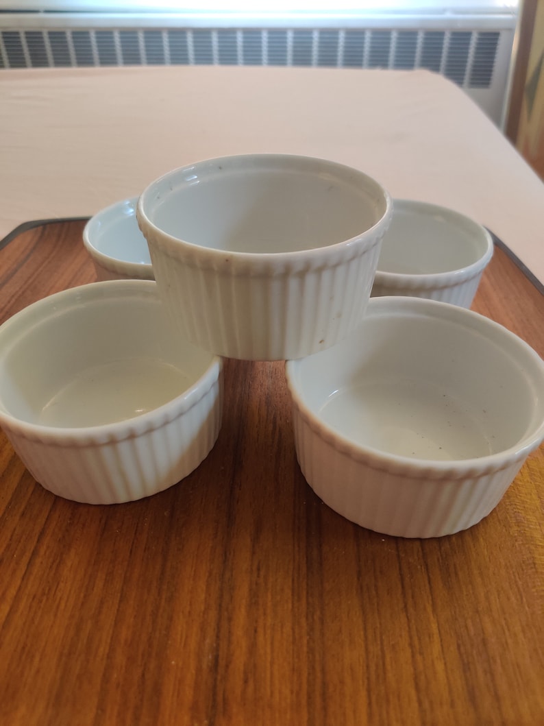 May include: A collection of five white, fluted ramekins arranged on a wooden cutting board. The ramekins are round with a classic design, perfect for baking and serving individual portions of desserts or savory dishes. The background includes a table and a window.